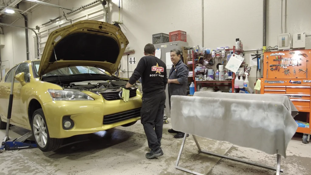 Two technicians inspecting the front of a vehicle with the hood open inside a Star Body Works collision repair shop.